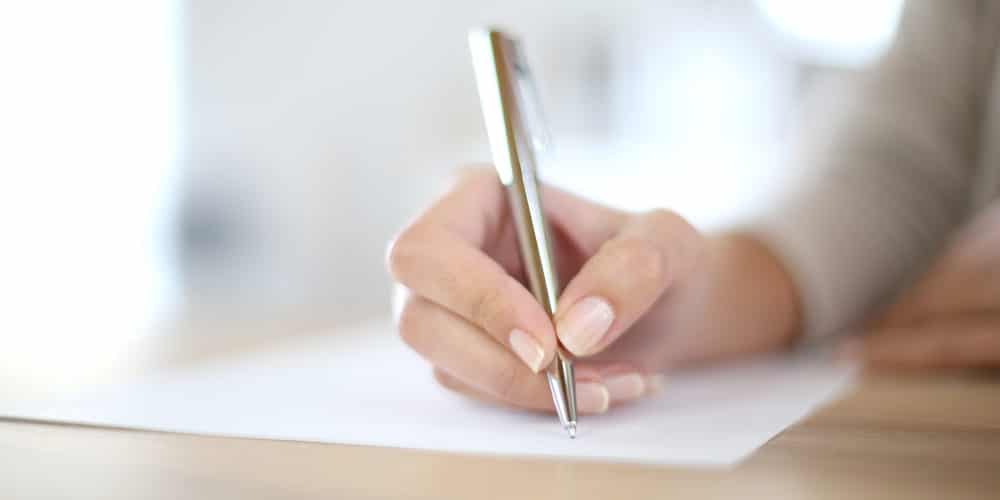 Mão feminina com unhas pintadas de branco assinando documento com caneta dourada