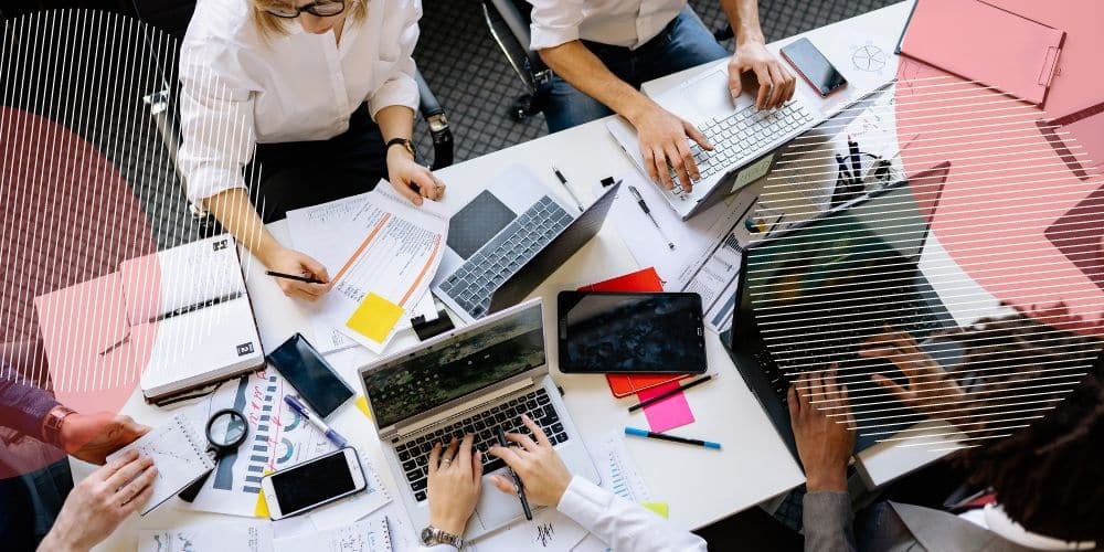 Vista superior de um conjunto de cinco profissionais trabalhando ao redor de uma mesa branca usando computadores, cadernos, documentos, celulares e tablets