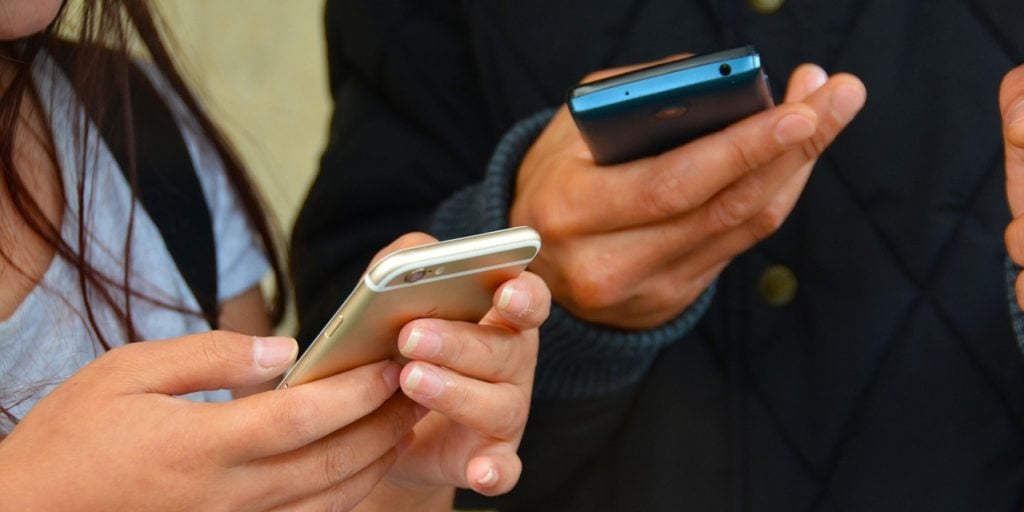 Homem e mulher mexem em seus smartphones lado a lado, a foto foca em suas mãos segurando os aparelhos.
