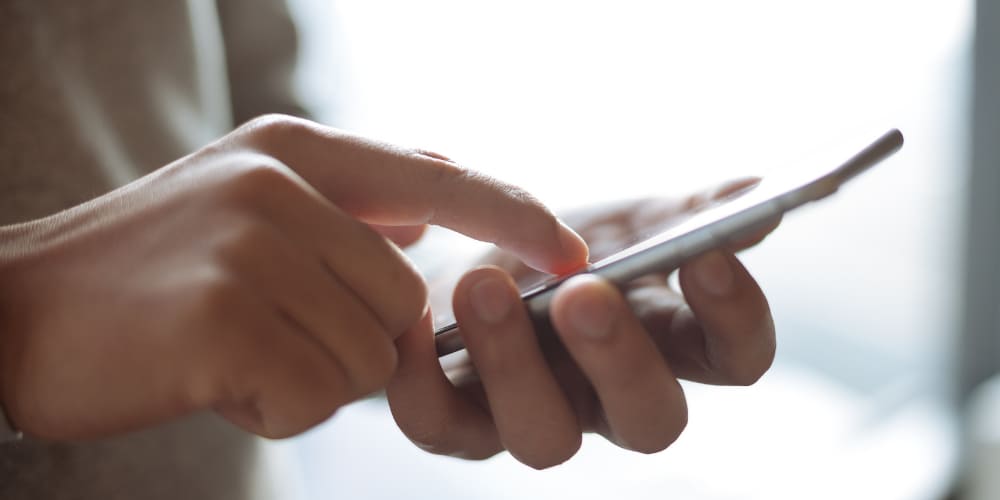 Mãos manuseando um smartphone em frente a uma janela ampla e clara