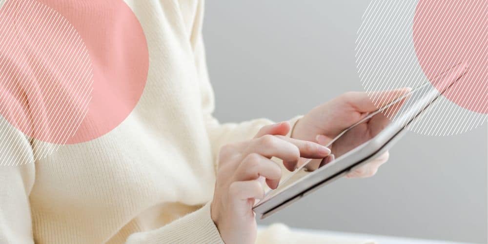 Foco nas mãos de uma mulher branca, vestida com uma blusa de lã off-white, mexendo em um tablet com a ponta dos dedos para controle ponto online gratuito