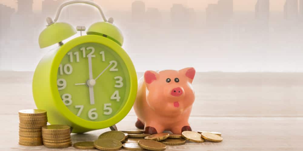 Relógio analógico verde-limão ao lado de um cofre em formato de cofrinho sobre mesa cheia de moedas