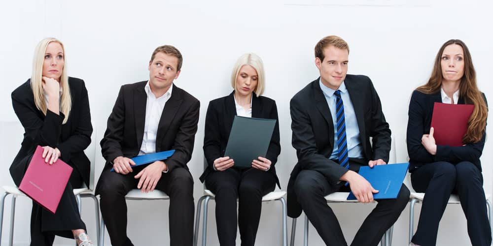 Cinco trabalhadores vestindo roupa social sentados lado a lado em corredor para um processo de seleção e recrutamento