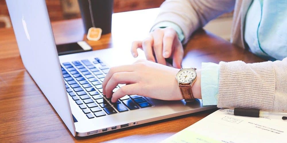 Mãos masculinas mexendo em um computador sobre mesa de madeira. O homem veste um relógio com pulseira de couro marrom e há uma xícara de chã ao lado dele sobre a mesa.