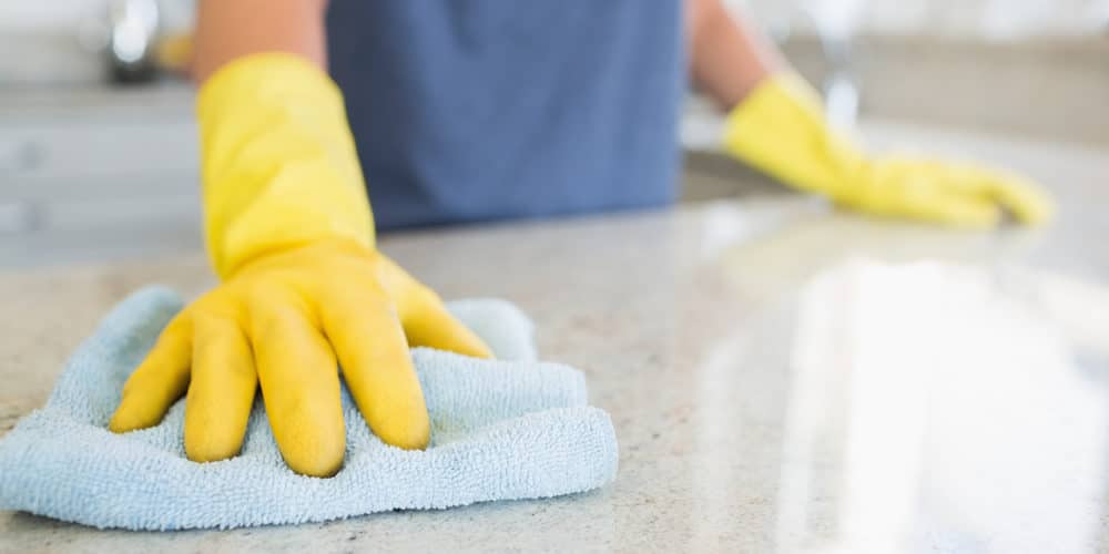 Pessoa com luvas amarelas feitas de borracha passando pano azul claro sobre bancada branca