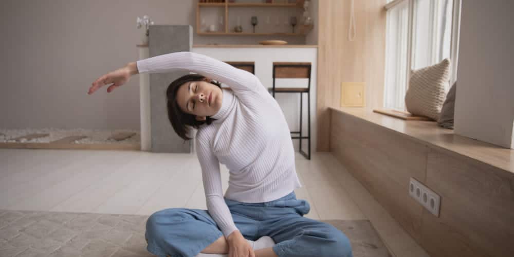 Mulher de cabelos escuros veste camiseta branca de manga longa e calças azuis claras confortáveis enquanto se alonga sentada no chão