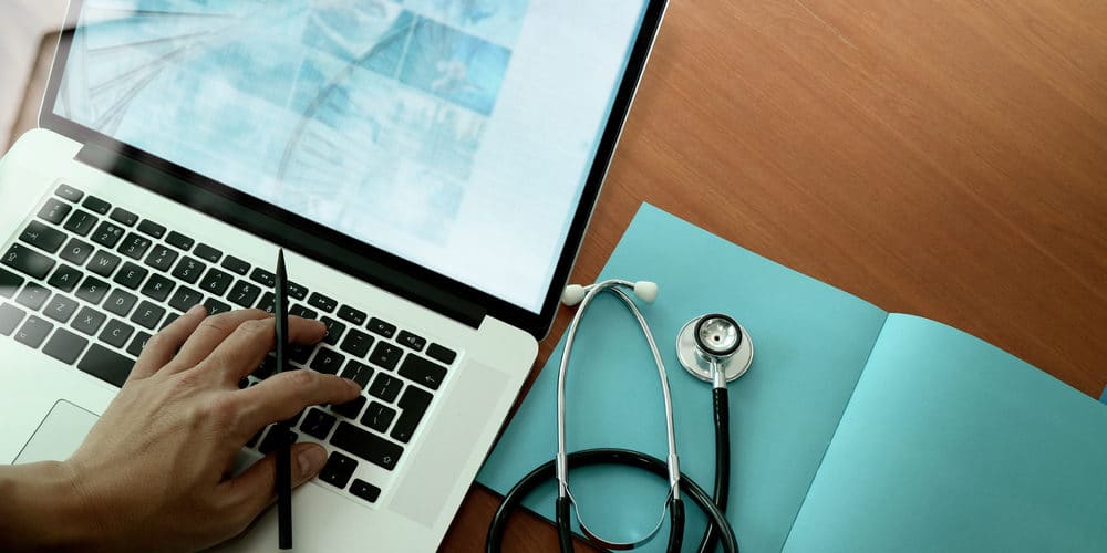 Médico mexendo em laptop sobre mesa de consultório. Ao lado do laptop há uma guia de receitas e um estetoscópio.