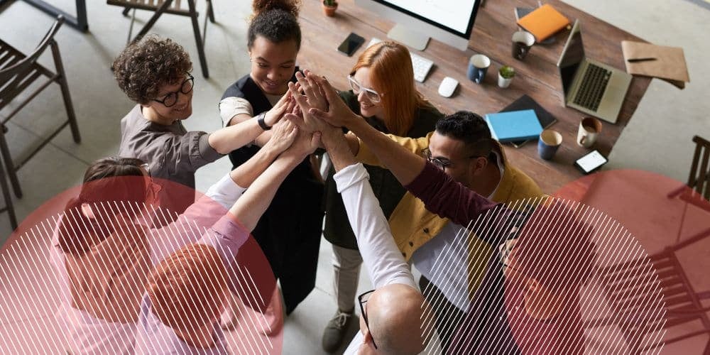 Grupo de 8 funcionários de diferentes gêneros e etnias erguendo as mãos e unindo-as em um grande cumprimento em escritório