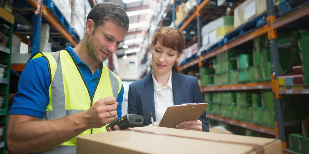 Homem vestindo uniforme para trabalho em estoque enquanto conversa com mulher que verifica informações em uma prancheta