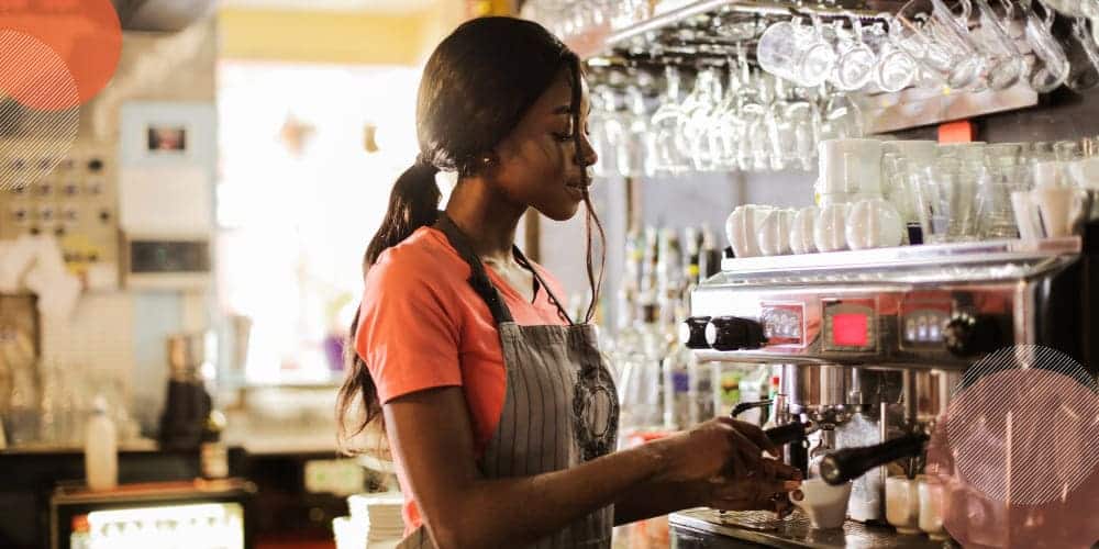 Mulher negra vestindo camiseta laranja e avental enquanto trabalha numa cafeteria