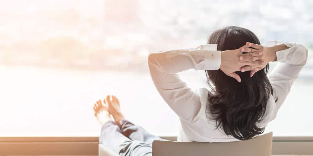 Mulher de cabelos castanhos sentada em cadeira de escritório, de costas para a foto, com os pés sobre mesa de madeira e mãos atrás da cabeça.