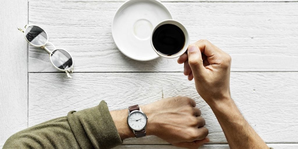 Vista de cima para baixo de uma pessoa checando as horas em seu relógio de pulso enquanto toma um café em uma xícara pequena