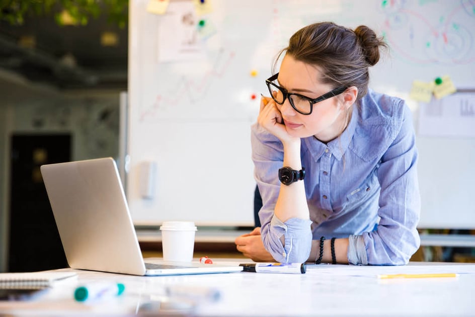 Mulher vestindo de camisa azul de mangas longas e usando óculos está apoiada sobre mesa de escritório enquanto olha para seu laptop