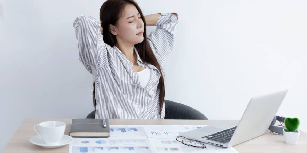 Mulher vestindo camisa social cinza estica os braços atrás da cabeça em frente à mesa de trabalho repleta de documentos e com um computador aberto em frente