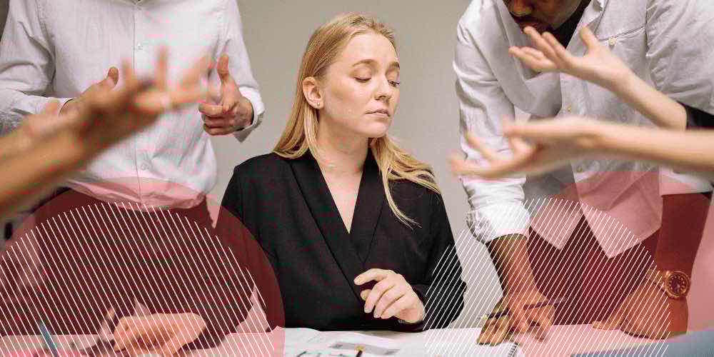 Mulher loira com roupas sociais sentada à mesa de escritório enquanto pessoas falam ao redor dela