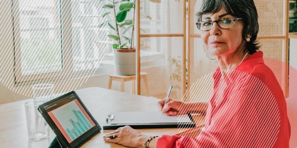 Mulher idosa com camisa rosada sentada em mesa de trabalho em frente ao computador