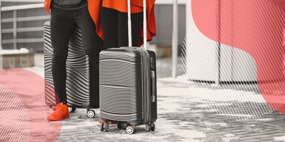 Duas pessoas vestidas com calça preta e casacos laranja esperam transporte até o aeroporto com suas bagagens de mão