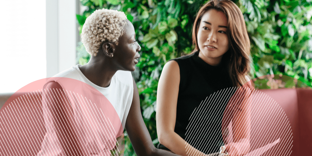 Mulher asiática vestida de preto conversa com uma mulher negra de cabelos loiros vestida de branco em um escritório