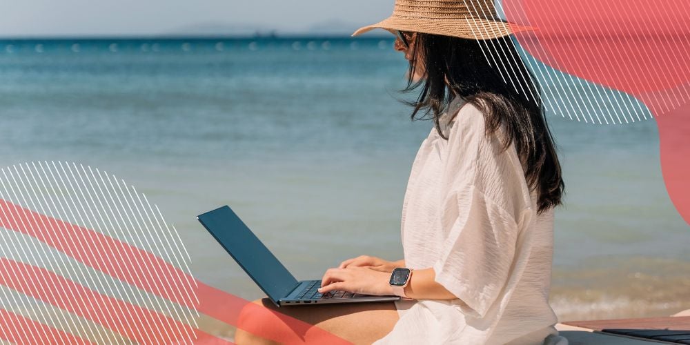 Mulher com camisa branca e chapeu de praia usando um laptop de frente para o mar