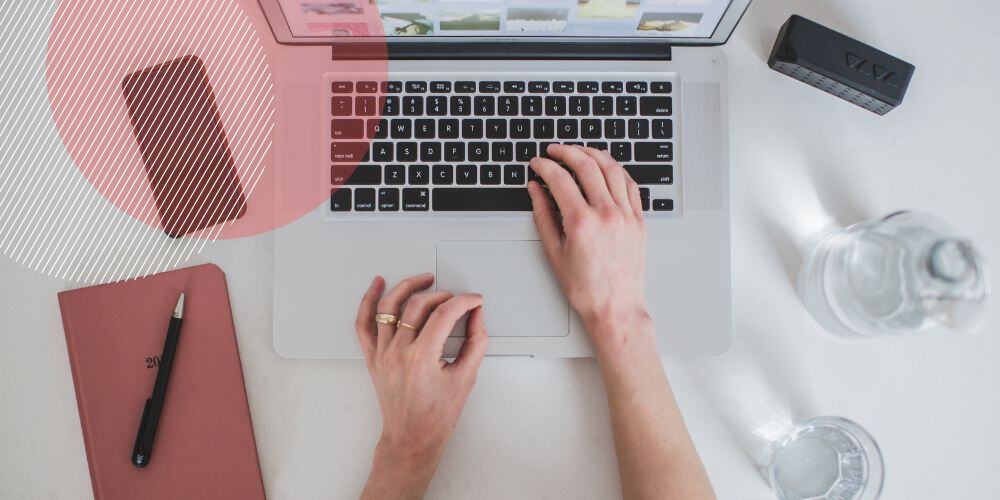 Mãos sobre teclas de laptop apoiado sobre mesa de trabalho branca com agenda e celular ao lado