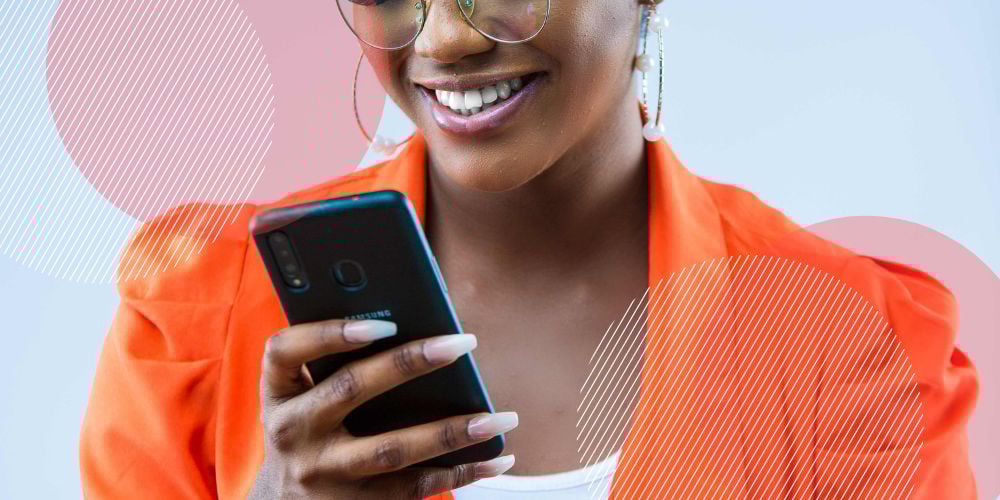 Mulher negra que veste um blazer laranja sorri enquanto mexe em seu smarphone