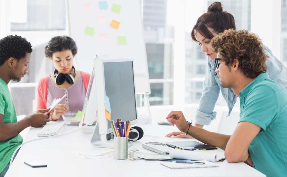 Quatro colegas reunidos em mesa de trabalho cheia de computadores, canetas, documentos e cadernos em escritório de decoração branca