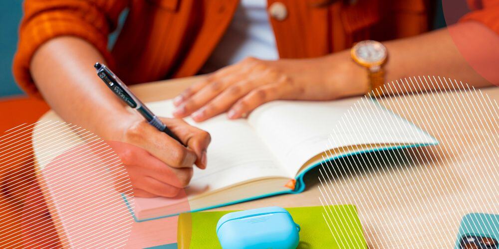 Pessoa vestida com blusa laranja faz anotação em caderno sobre mesa de escritório cheia de mateiriais coloridos