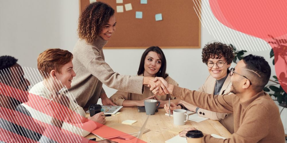 Grupo de colegas de trabalho reunidos ao redor de mesa de escritório enquanto dois deles apertam as mãos e riem