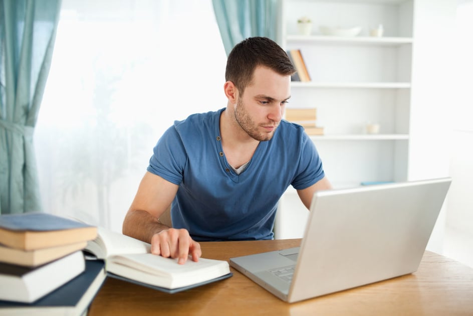Homem branco de cabelos castanhos e curtos vestido com camiseta azul trabalha remotamente em sua casa sobre uma mesa de madeira que suporta laptop, telefone e livros