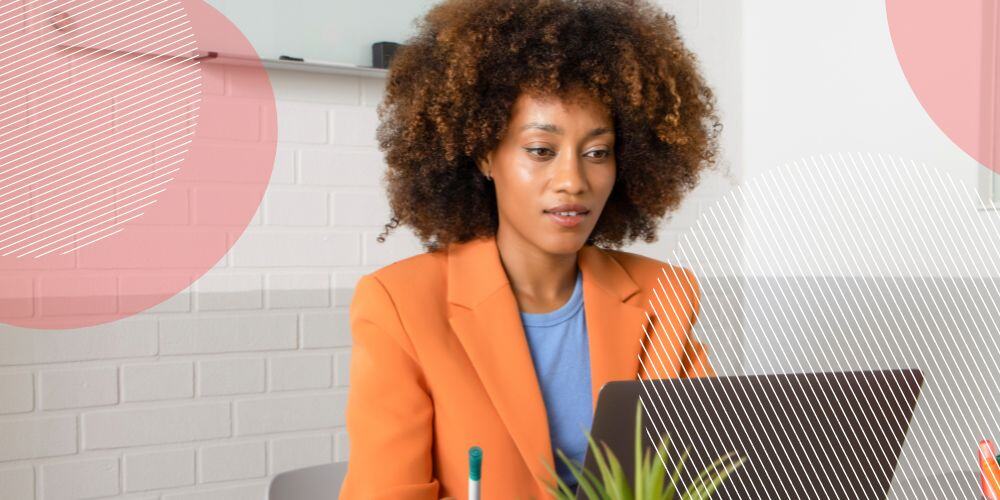 Mulher negra vestindo blazer laranja sentada em frente à mesa de trabalho enquanto escreve em seu computador