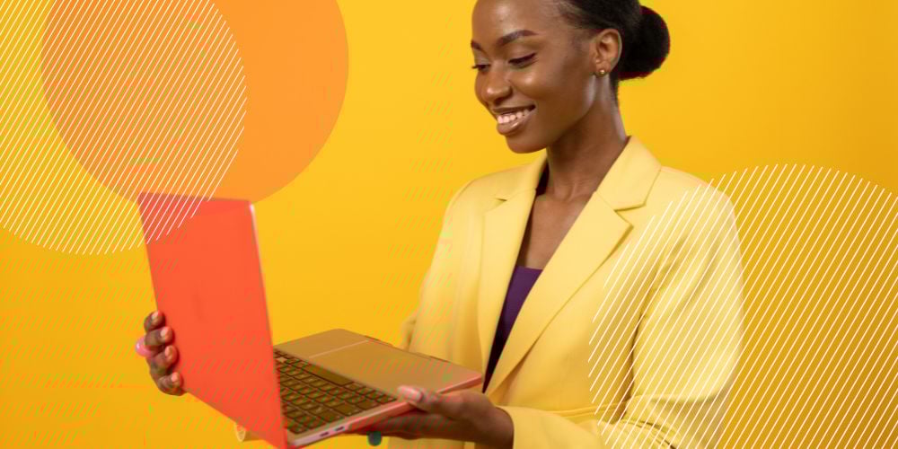 Mulher negra vestida com terno amarelo segura computador laranja nas mãos enquanto sorri